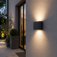 Modern outdoor wall light fixture on a house exterior with plants and glass door in the background.