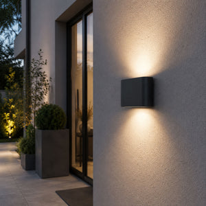 Modern outdoor wall light fixture on a house exterior with plants and glass door in the background.