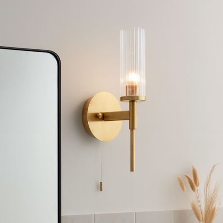 Gold wall sconce with clear glass shade on a light tiled wall next to a mirror and basket.